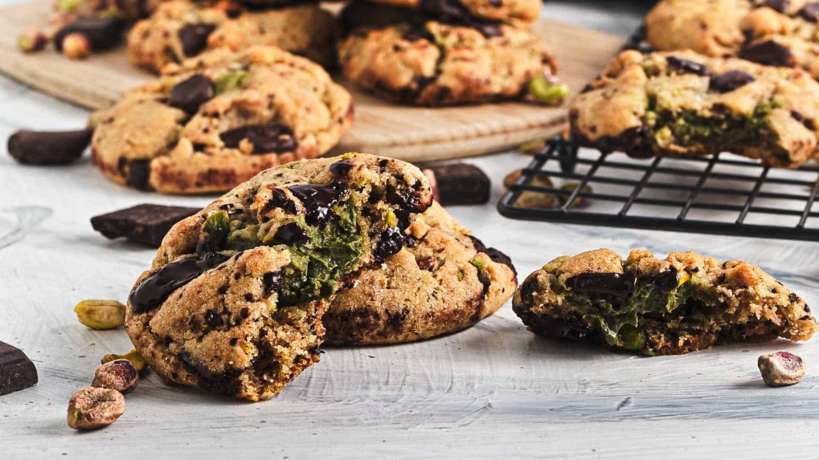 A close-up of a chunky cookie with chocolate pieces and pistachios on a wooden board.