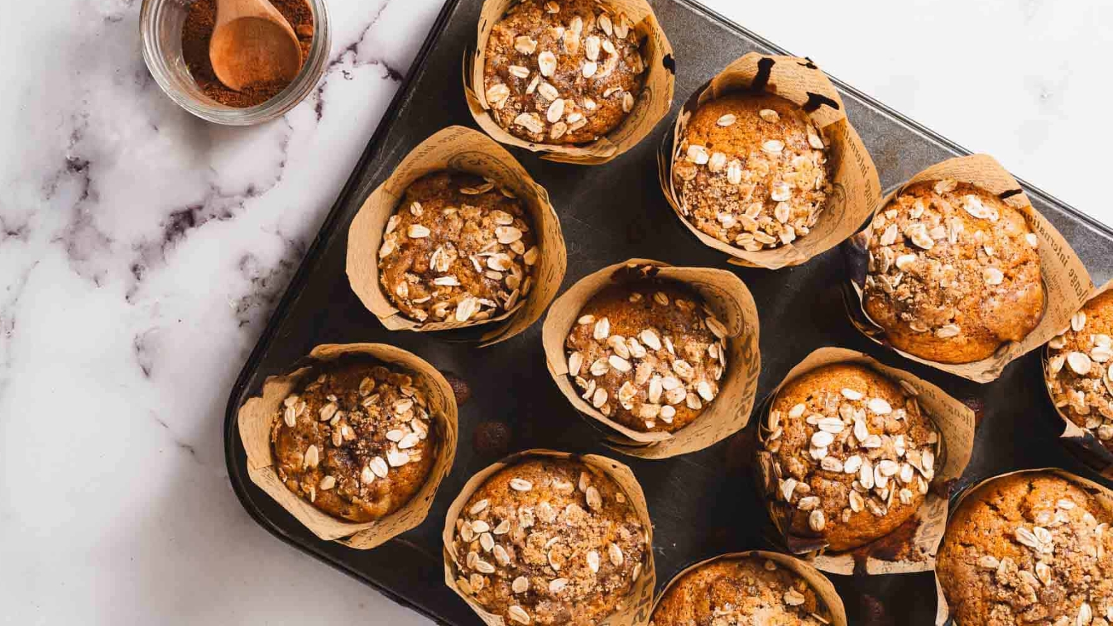 A muffin topped with oats is placed on a decorative paper wrapper. The muffin sits on a marble surface, with crumbs scattered around it.