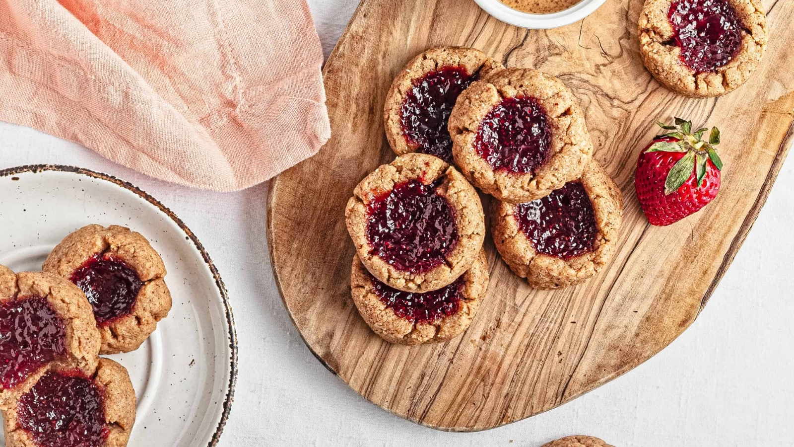 Thumbprint cookies with strawberry jam on a baking tray, surrounded by fresh strawberries.