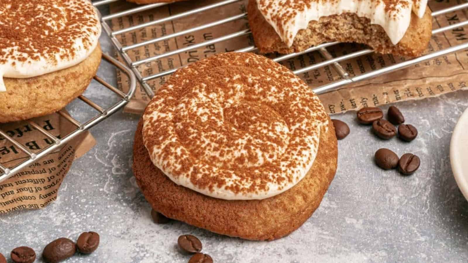 A frosted cookie dusted with cocoa powder, surrounded by coffee beans and baked cookies on a cooling rack.