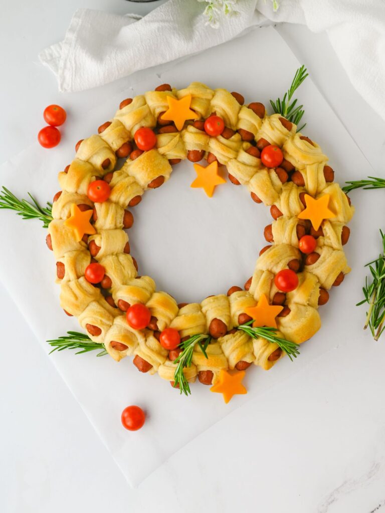 Pigs in a Blanket Christmas Wreath Appetizer with cheese stars, rosemary, and cherry tomatoes.