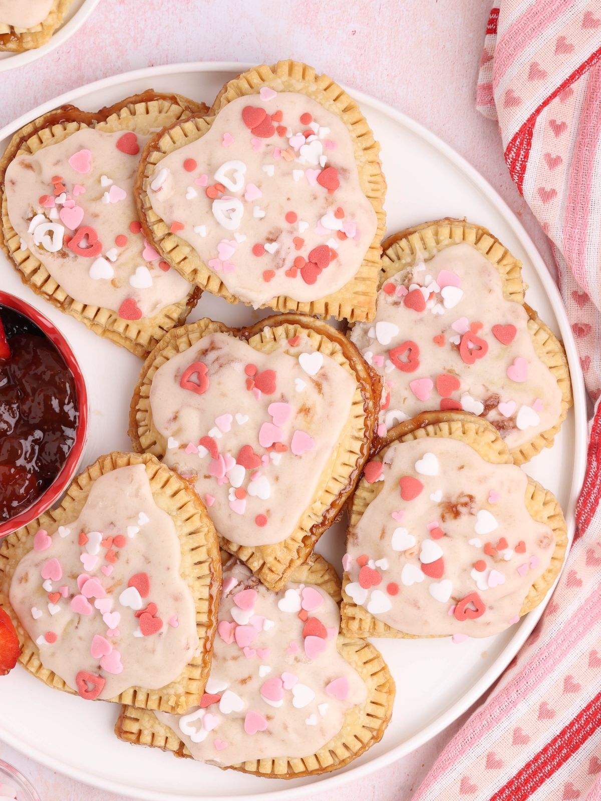 Heart-shaped strawberry hand pies topped with vanilla glaze and pink and red sprinkles on a white plate.