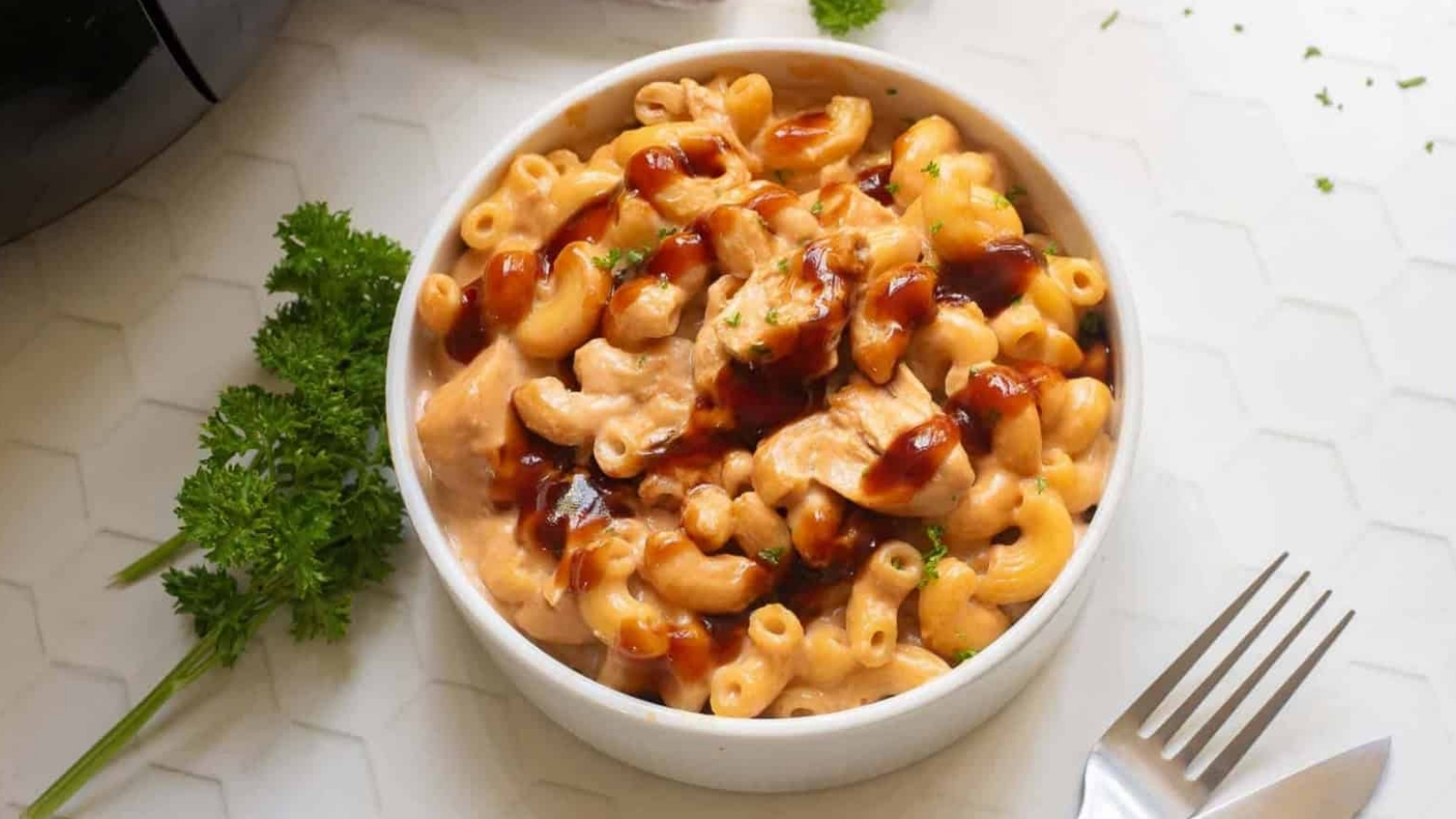A bowl of macaroni and cheese with pieces of chicken, topped with a drizzle of brown sauce, sits on a white surface beside a fork and sprigs of parsley.