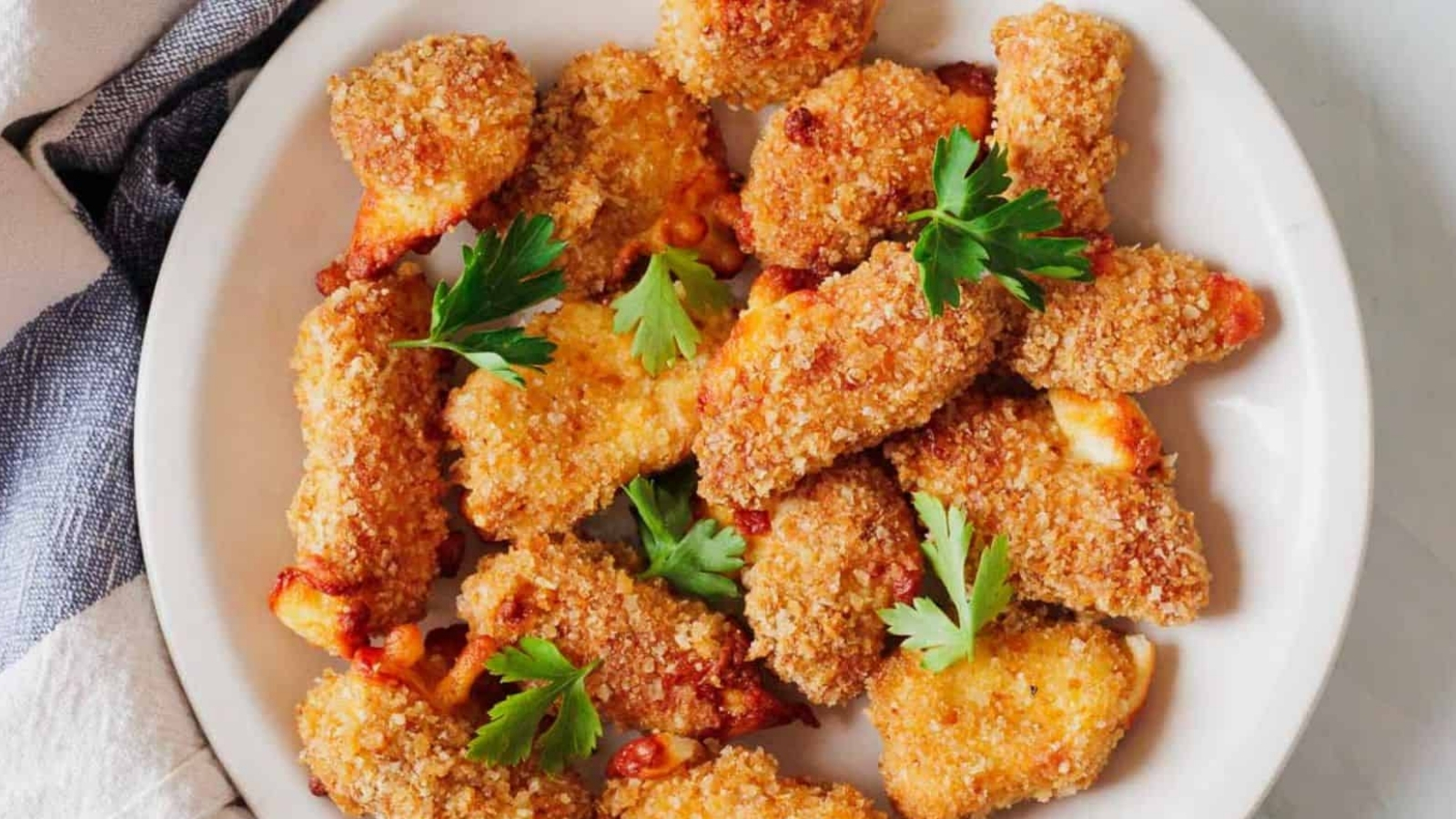 A plate of breaded chicken nuggets garnished with parsley. The nuggets are golden brown, and the dish is placed on a white surface with a striped cloth partially visible.