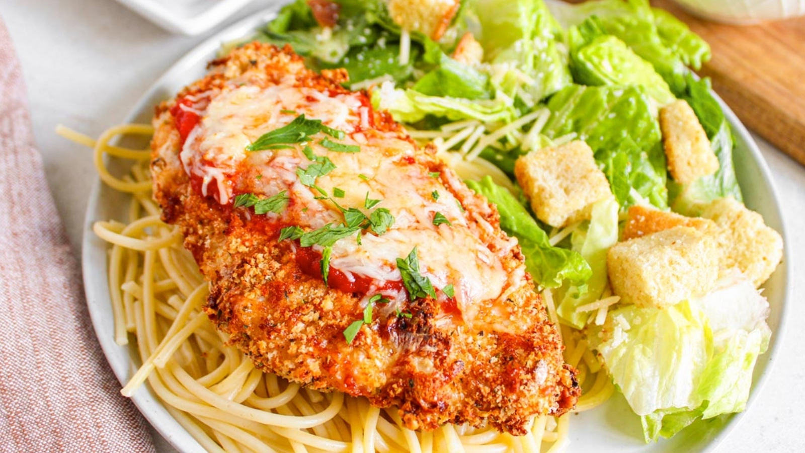 A plate with breaded chicken topped with tomato sauce and cheese on spaghetti, served with a green salad and croutons.