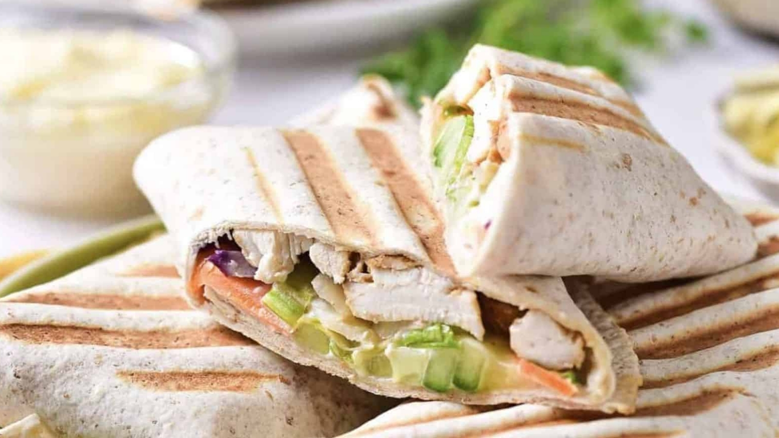 Two grilled wraps are cut in half, showing fillings of grilled chicken, lettuce, cucumber, and other vegetables. The wraps have visible grill marks and are stacked on a plate with a blurred background.