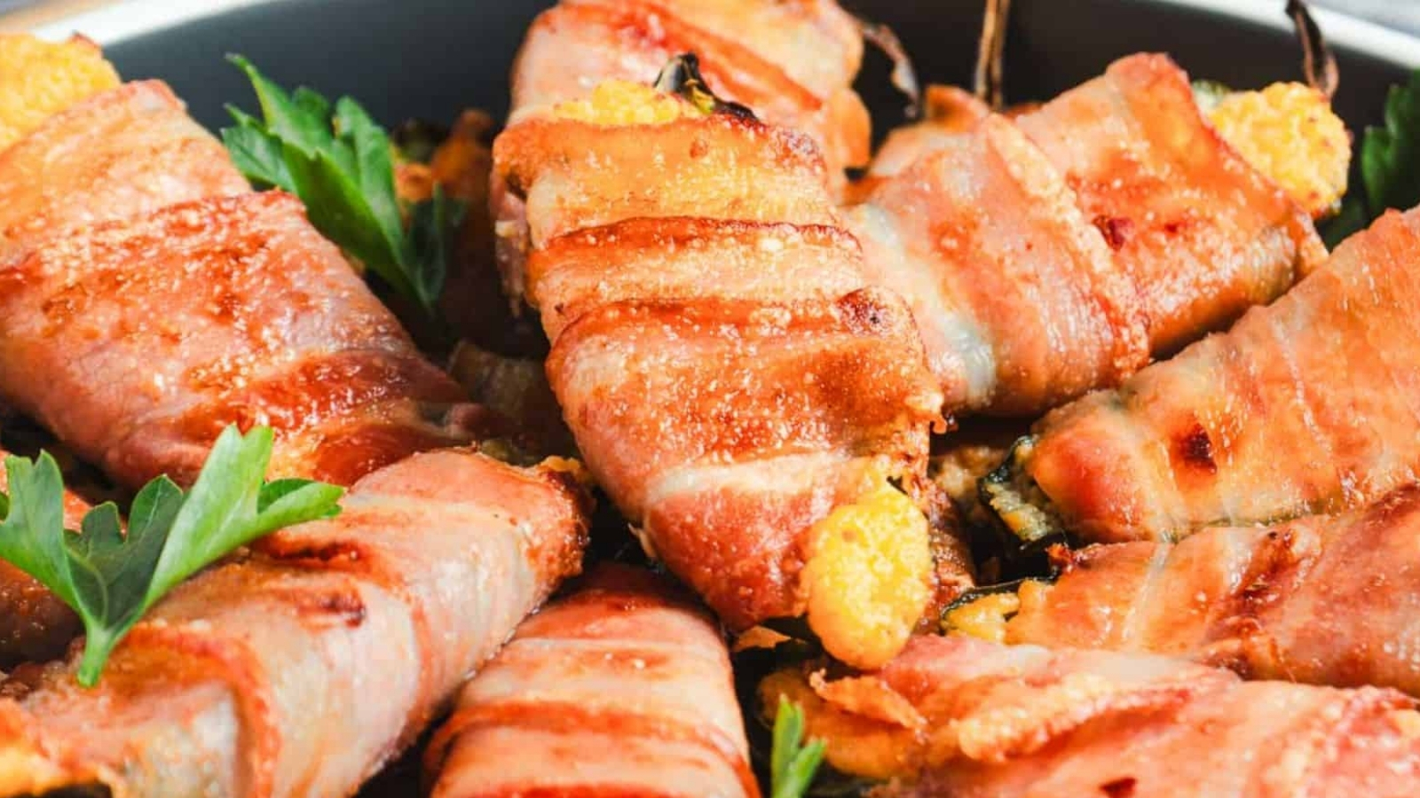Plate of bacon-wrapped peppers filled with cheese, garnished with parsley, next to a grated cheese block and grater on a wooden board.