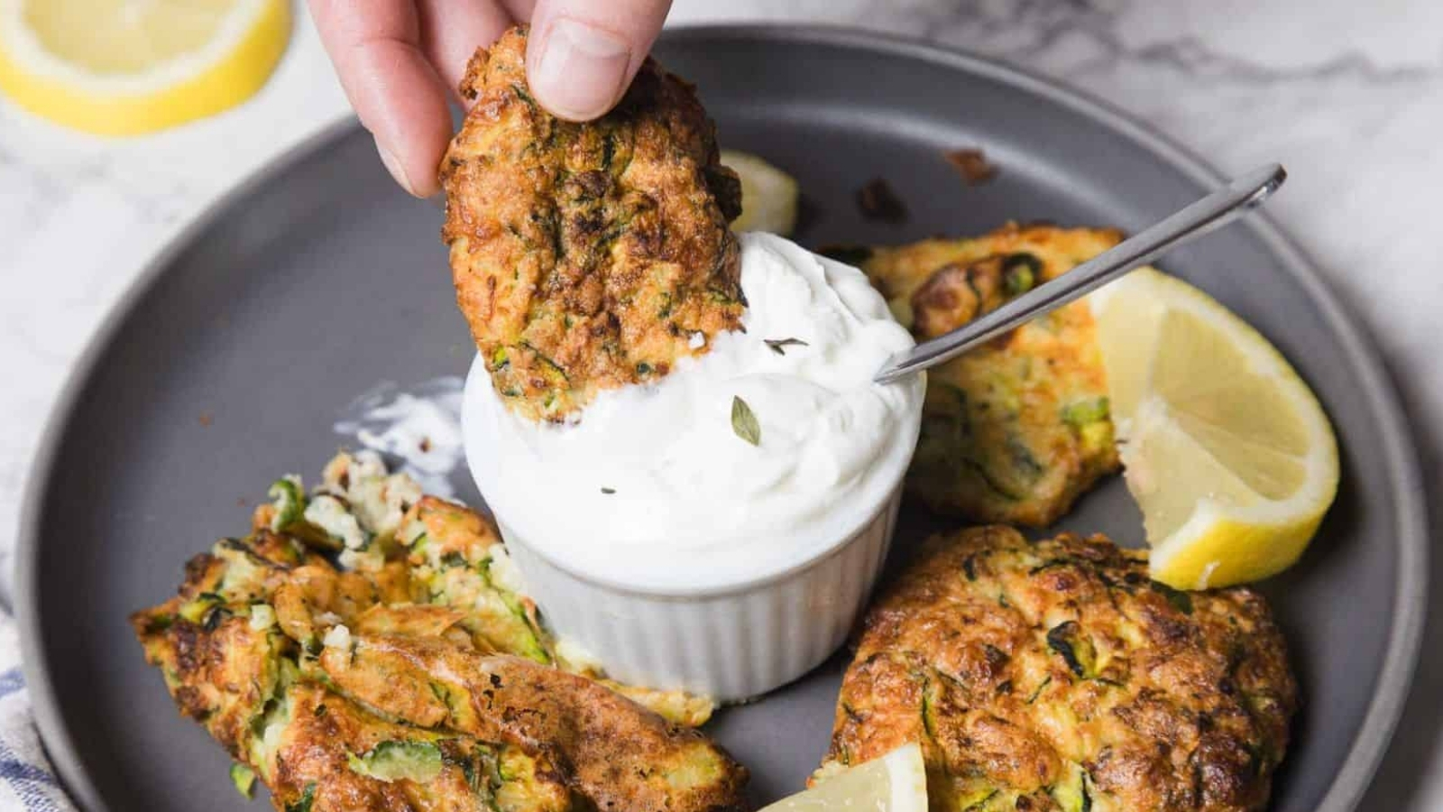 A hand dips a fried zucchini fritter into a small bowl of white cream sauce on a gray plate. Other fritters and lemon wedges are arranged around the bowl on a marble surface.