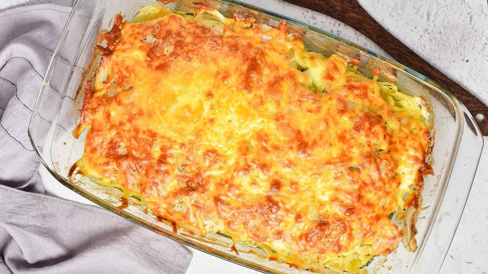 A rectangular glass baking dish filled with a baked casserole topped with melted, browned cheese sits on a white surface beside a folded gray cloth and a wooden utensil.