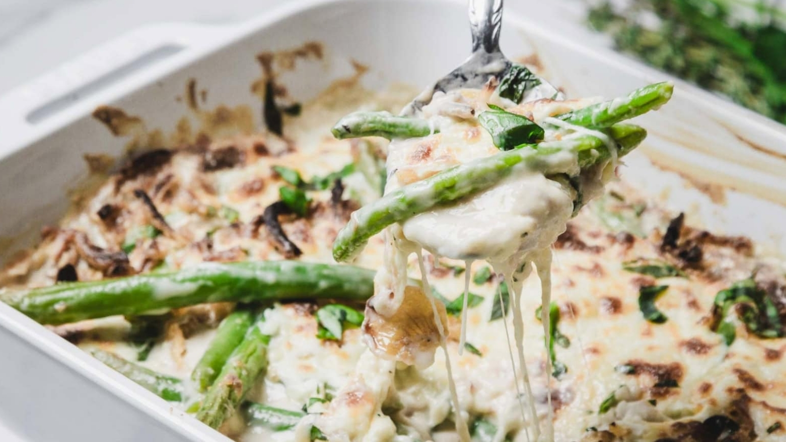 A cheesy green bean casserole being served from a white baking dish with a spoon.