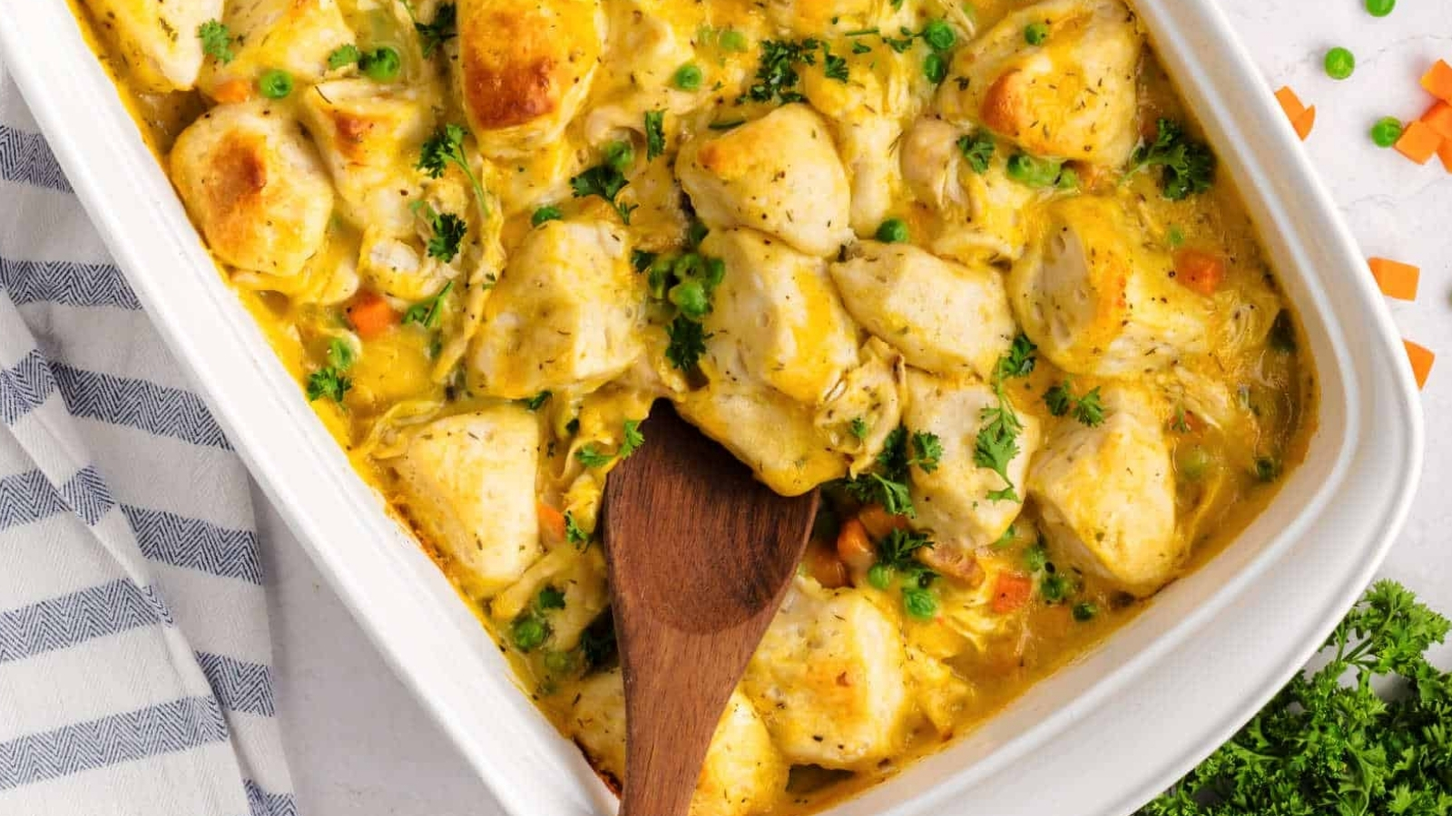 A casserole dish filled with baked chicken, mixed vegetables, and creamy sauce, with a wooden spoon resting inside.