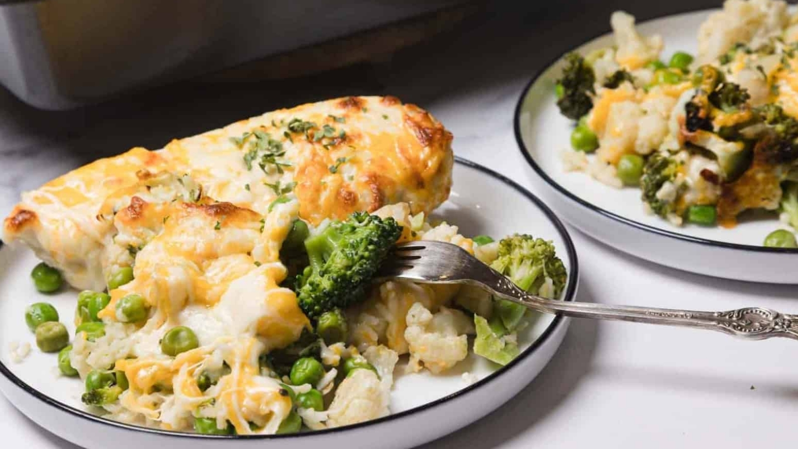 A plate of cheesy vegetable casserole with broccoli, peas, and cauliflower, with a fork lifting a bite.