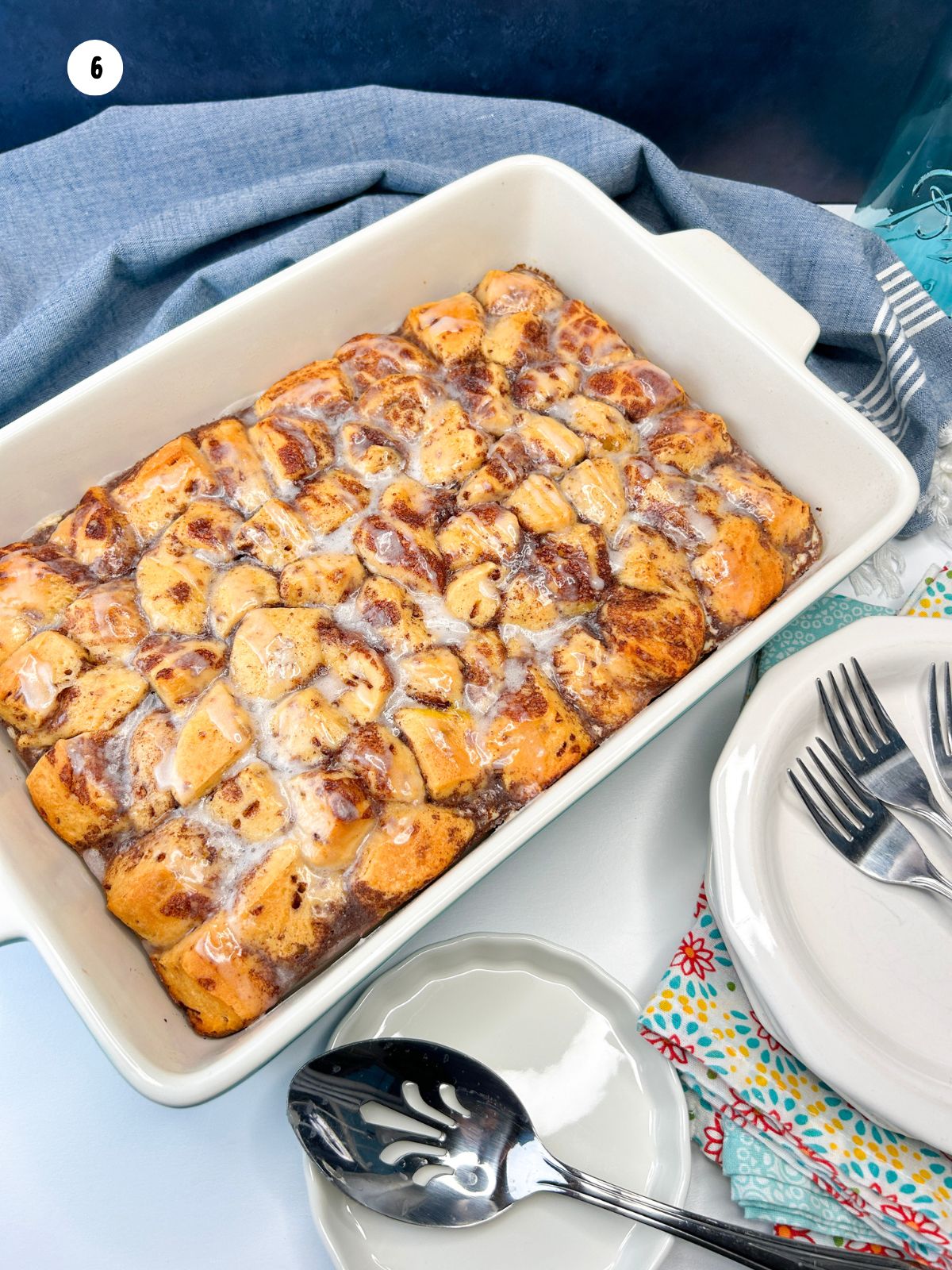 baked casserole with Pillsbury cinnamon rolls with frosting ready to serve in casserole dish with serving dishes and utensils nearby.