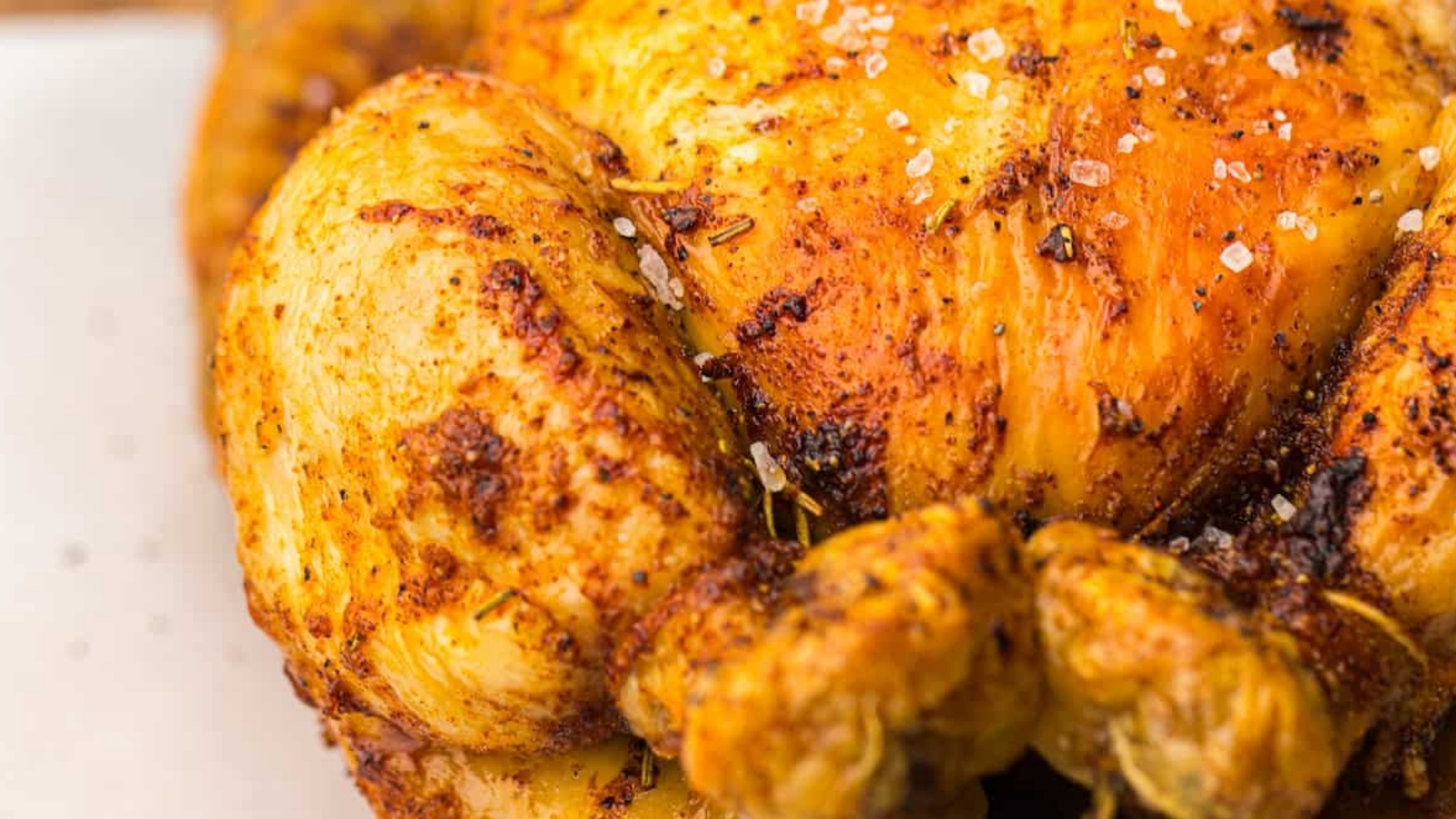 Close-up of a roasted chicken with crispy, golden-brown skin seasoned with herbs and coarse salt, on a wooden cutting board.
