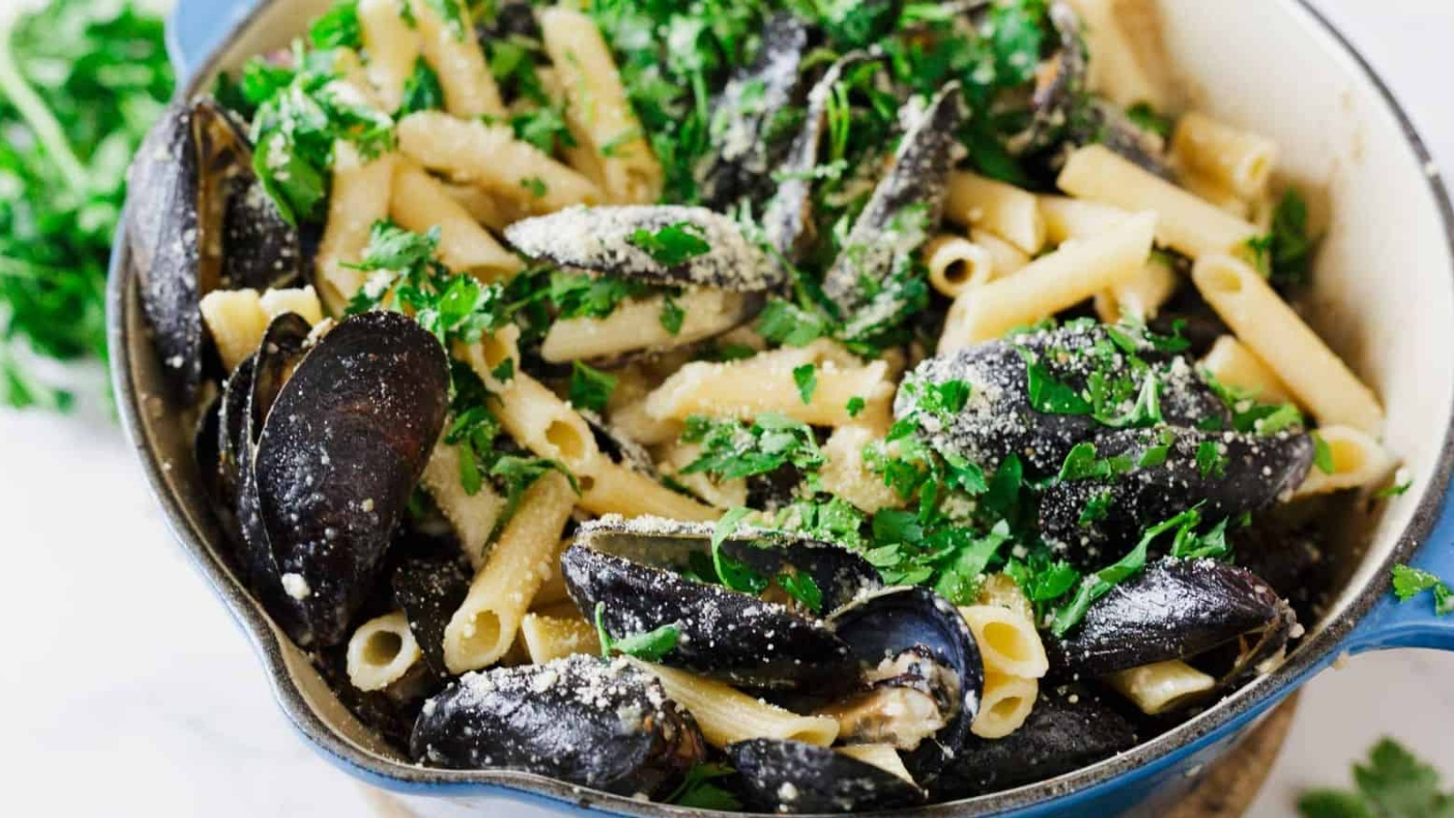 A close-up of a blue pot filled with cooked penne pasta, mussels in shells, chopped parsley, and grated cheese, with some fresh parsley scattered around the pot on a white surface.