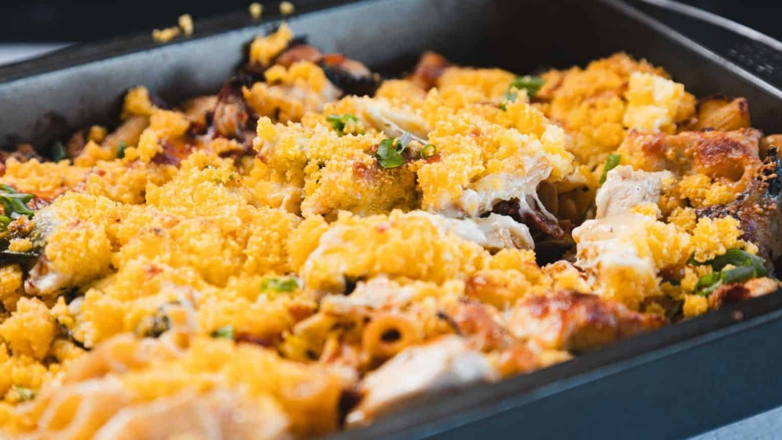 A close-up of a baked pasta dish in a rectangular metal pan with a golden breadcrumb topping, pieces of pasta, baked cheese, and green herbs visible.