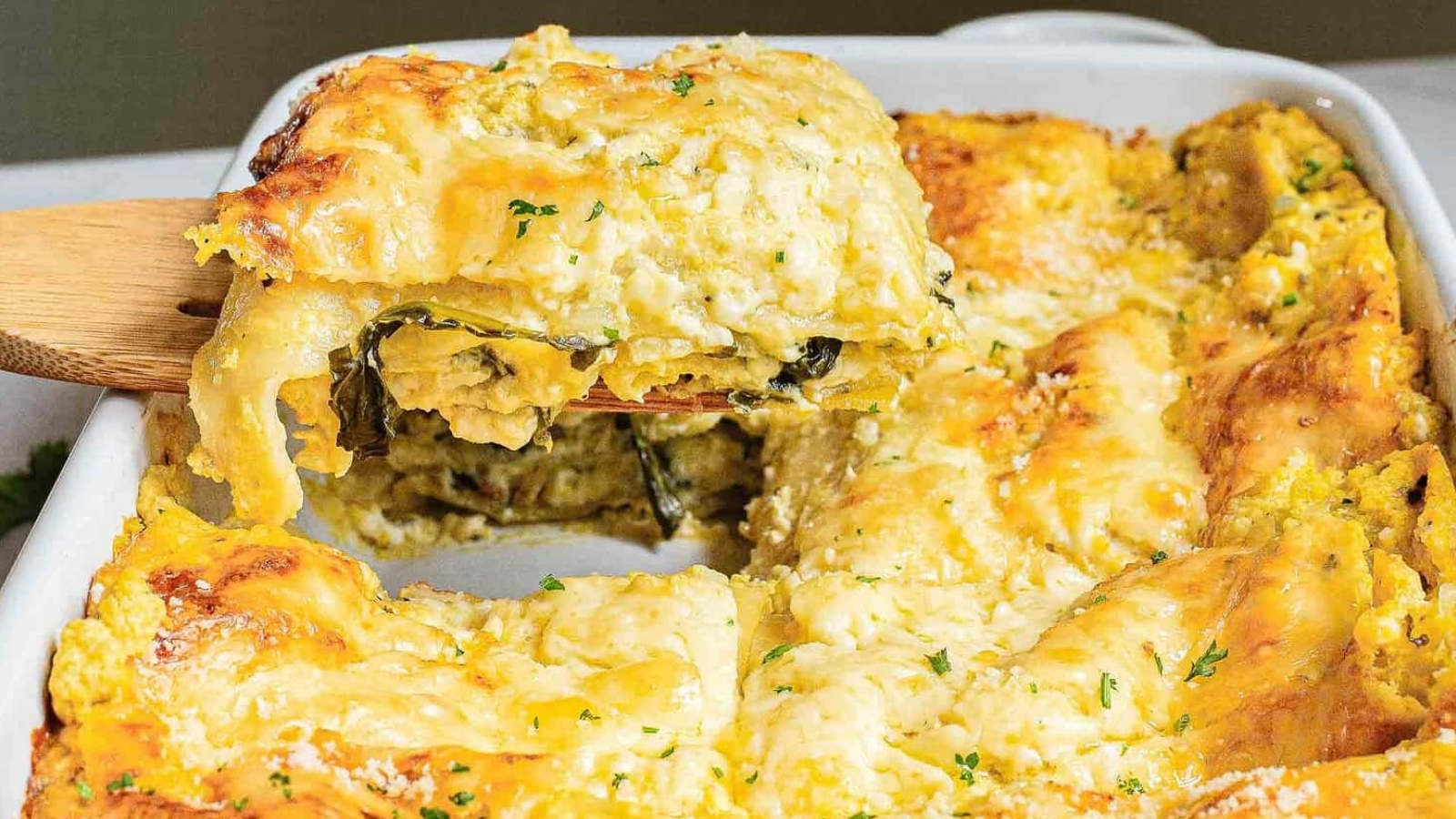 A baked dish of zucchini lasagna with a golden-brown cheese topping. A wooden spatula lifts a square piece, revealing layers of zucchini, cheese, and spinach. The casserole dish is partly cut, showing more servings. Garnished with herbs.