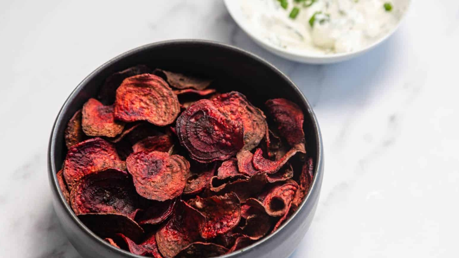 A black bowl filled with dark red beet chips sits on a white surface. In the background, there is a small white bowl containing a creamy dip with green herbs.