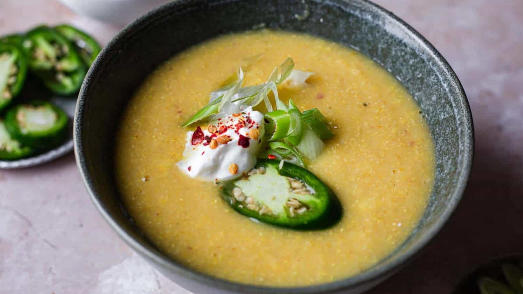 A bowl filled with yellow corn soup topped with a dollop of white cream, sliced green chili, and chopped green onion.