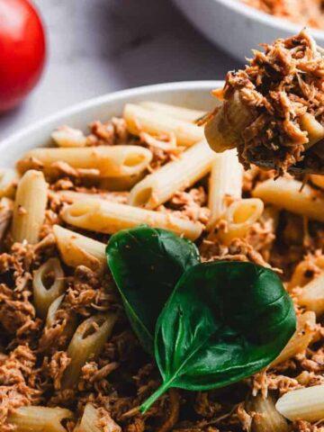 A bowl of penne pasta topped with shredded meat and garnished with fresh basil leaves. A fork is placed in the bowl. Red tomatoes and additional fresh basil leaves are on the marble surface nearby.