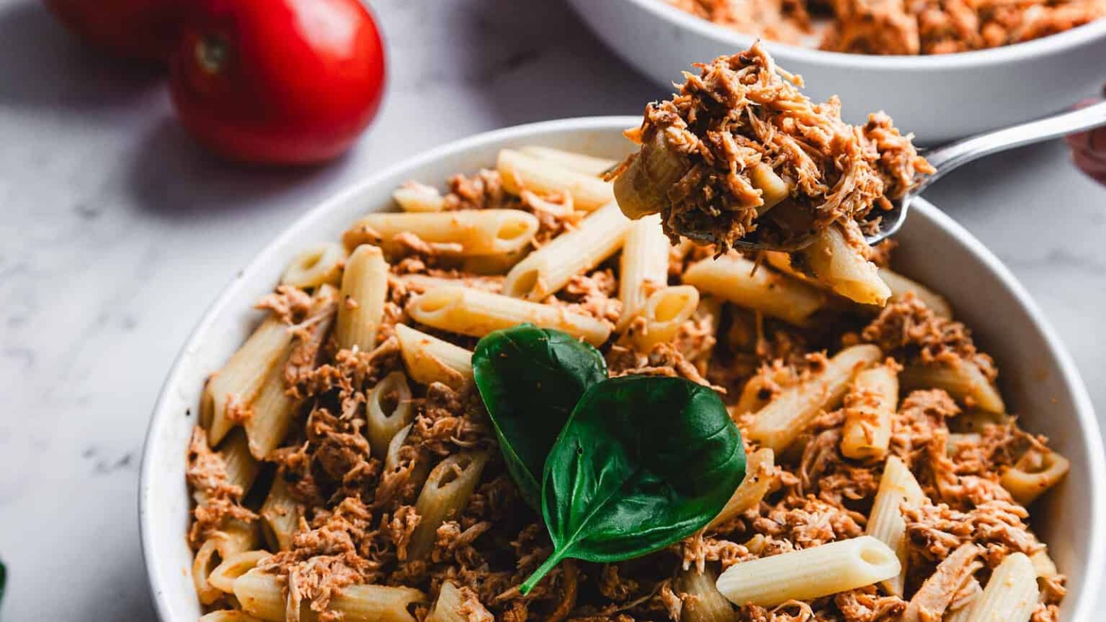 A bowl of penne pasta topped with shredded meat and garnished with fresh basil leaves. A fork is placed in the bowl. Red tomatoes and additional fresh basil leaves are on the marble surface nearby.
