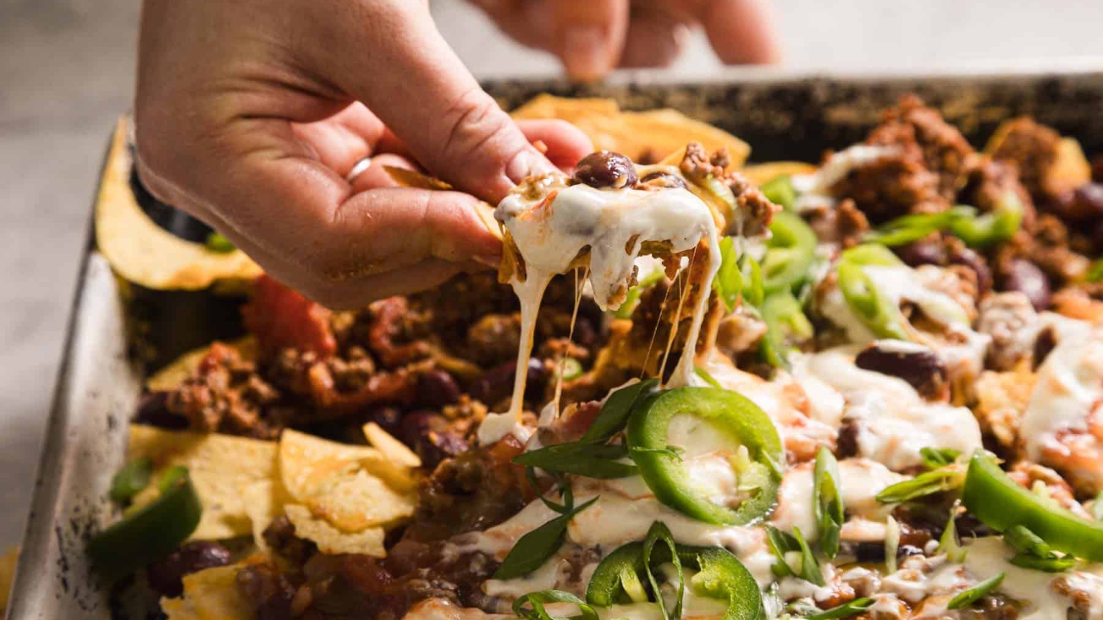 A close-up of nachos topped with melted cheese, ground beef, sliced jalapeños, chopped green onions, sour cream, and beans. The tortilla chips are spread out with the toppings generously applied, creating a colorful and appetizing presentation.