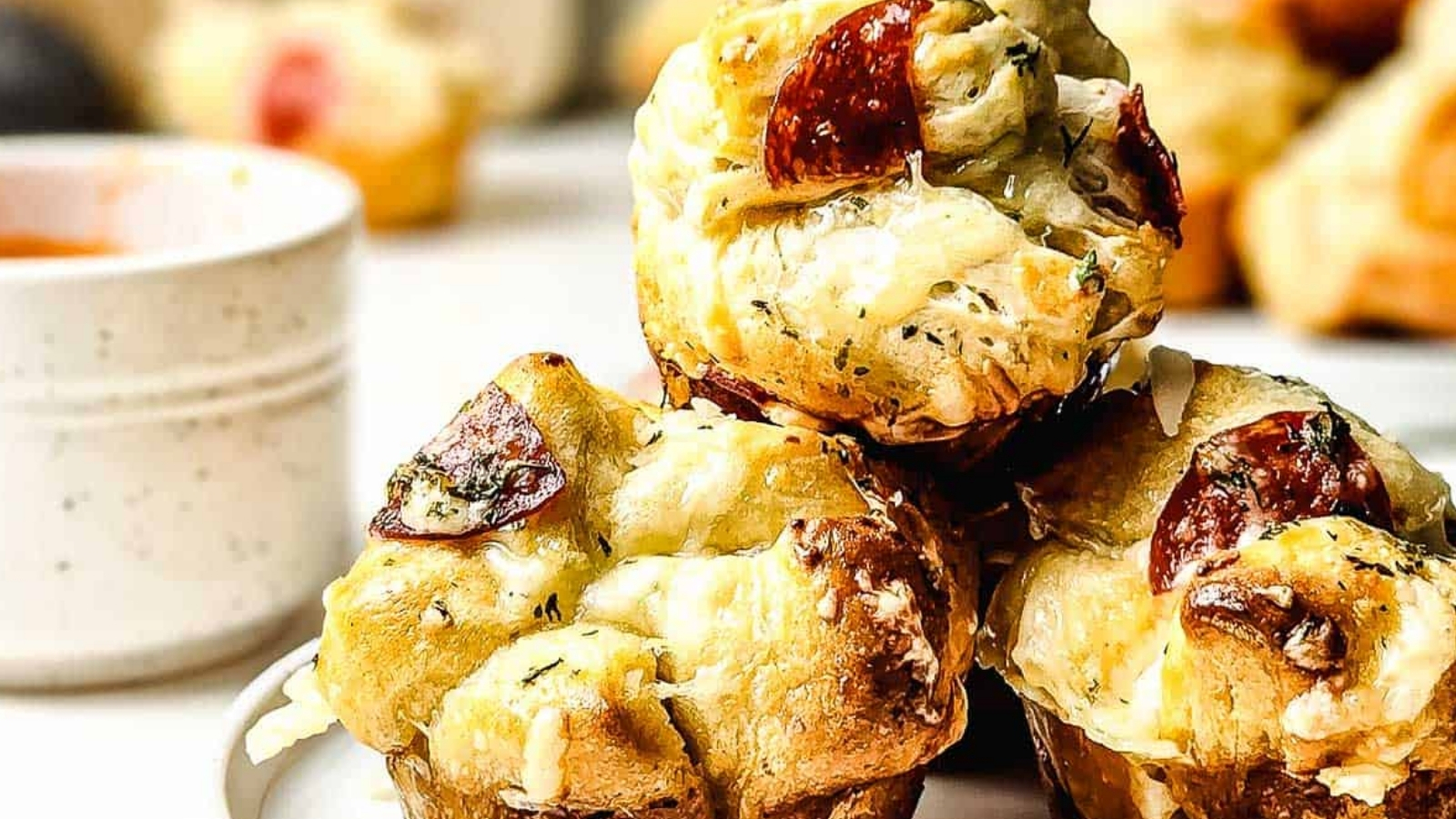 Three muffin-shaped pieces of pull-apart bread with melted cheese and pepperoni are stacked on a plate. A small bowl of dip and more bread pieces are visible in the background.