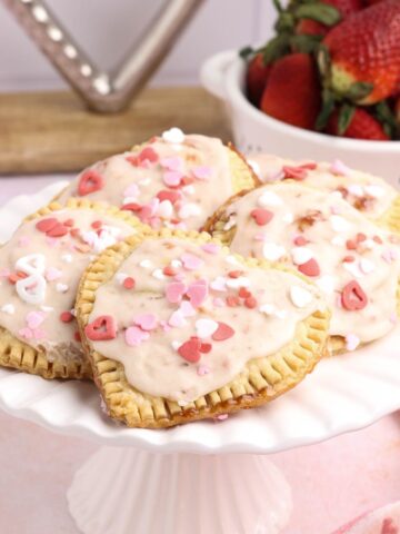 Strawberry filled heart shaped hand pies topped with vanilla glaze and pink and white sprinkles on a white cake stand.