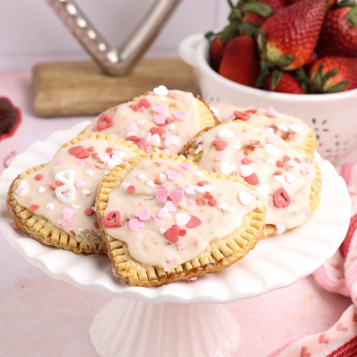 Strawberry filled heart shaped hand pies topped with vanilla glaze and pink and white sprinkles on a white cake stand.