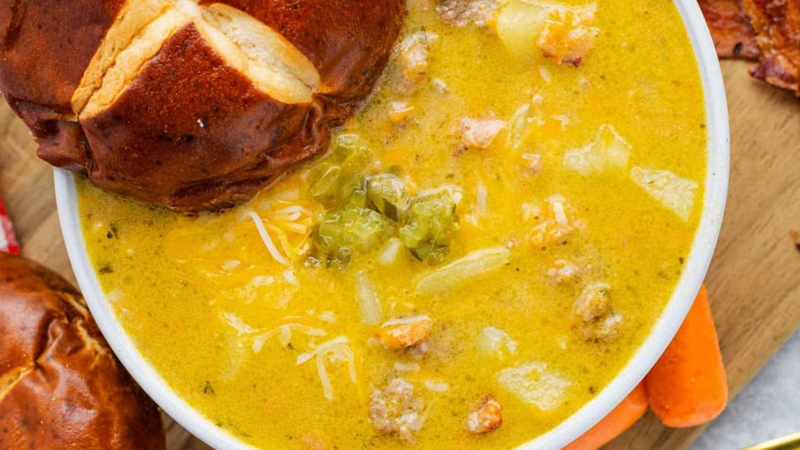 A bowl of cheesy soup with ground meat, potatoes, and vegetables, served with a pretzel bun.