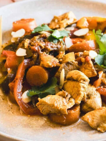 A plate of chicken stir-fry with sliced carrots, bell peppers, and mushrooms, garnished with cilantro and almond slices. A fork rests on the plate, and a yellow cloth is visible in the background.