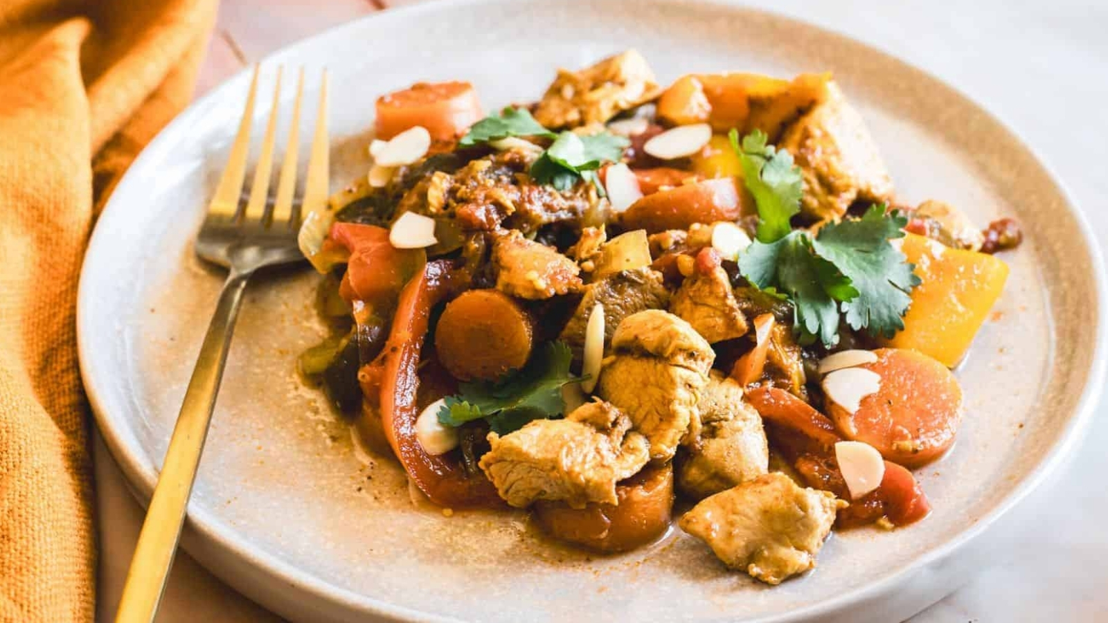 A plate of chicken stir-fry with sliced carrots, bell peppers, and mushrooms, garnished with cilantro and almond slices. A fork rests on the plate, and a yellow cloth is visible in the background.