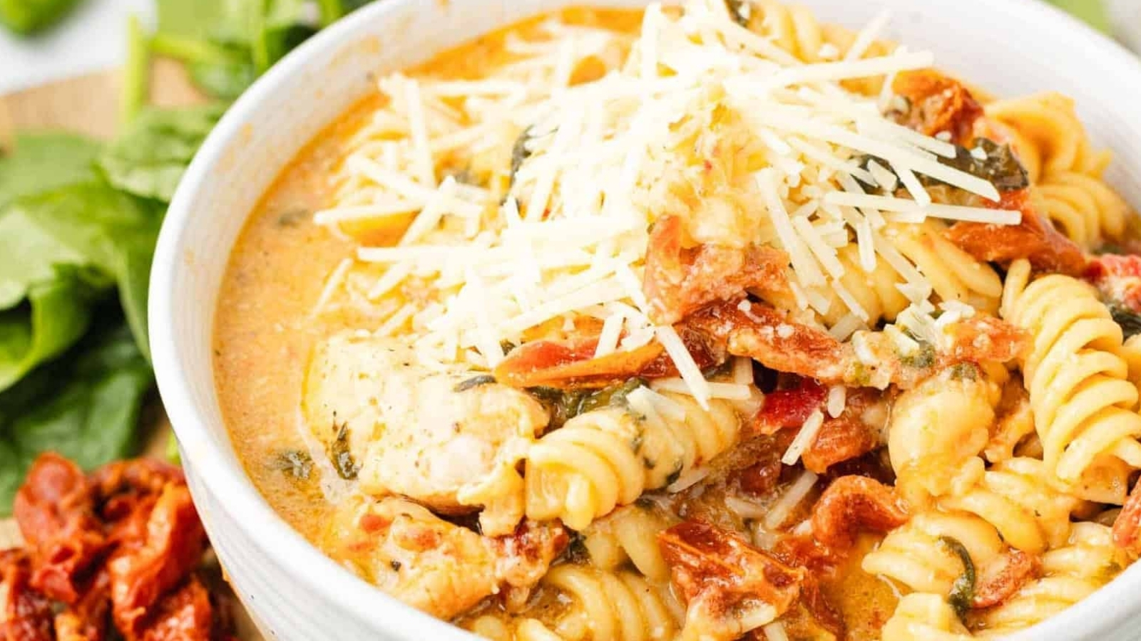 A bowl of creamy pasta with chicken, sun-dried tomatoes, spinach, and shredded cheese on top.