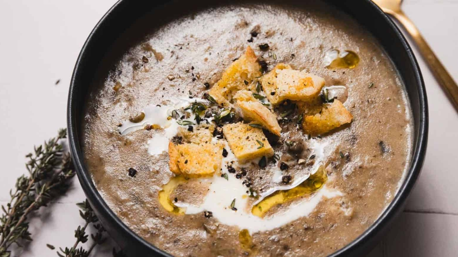 A bowl of creamy mushroom soup garnished with croutons, a drizzle of olive oil, and fresh herbs. A golden spoon lies nearby, and sprigs of thyme are placed on the table beside the bowl.