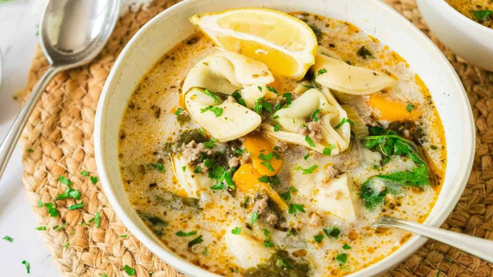 A bowl of creamy tortellini soup with spinach, lemon wedges, and herbs, served with a spoon.