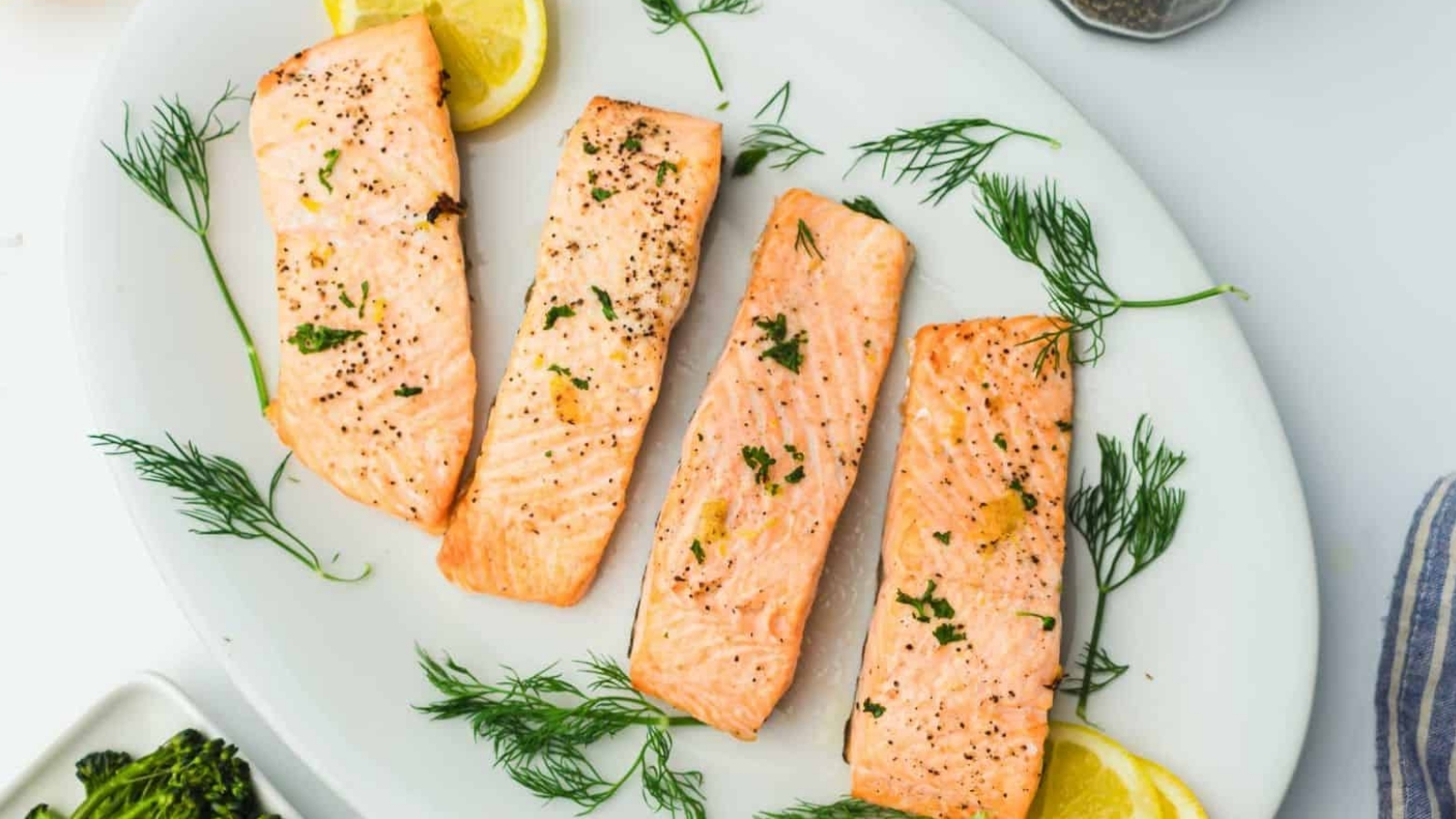 Four grilled salmon fillets garnished with fresh dill and lemon slices are arranged on a white oval platter. The fillets are seasoned with herbs and pepper. The edge of a striped cloth and a serving of broccoli are partially visible.