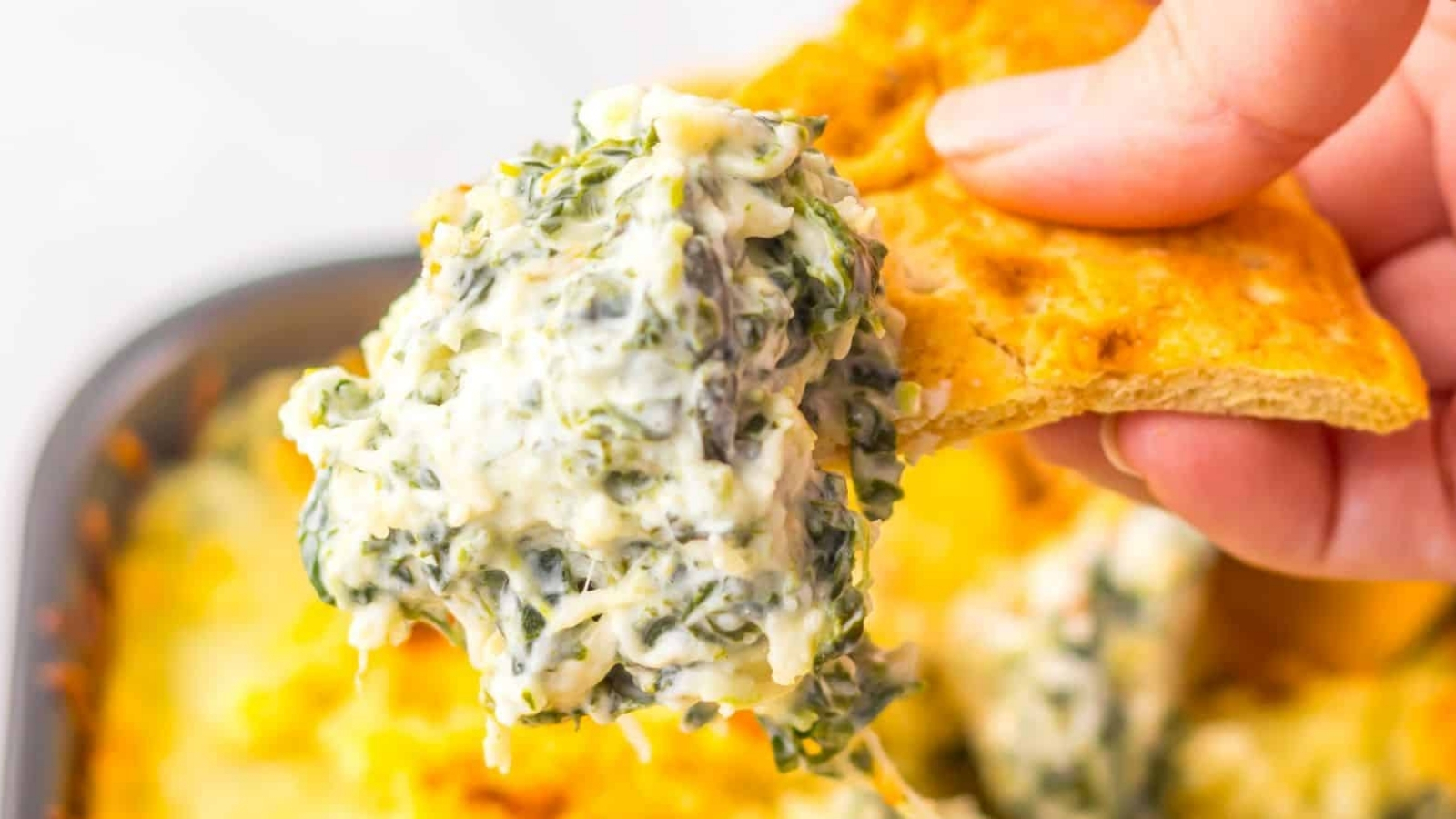A close-up of spinach artichoke dip on a cracker held by a hand. The dip is creamy with visible green spinach and bits of artichoke. The background shows a baking dish filled with the dip.