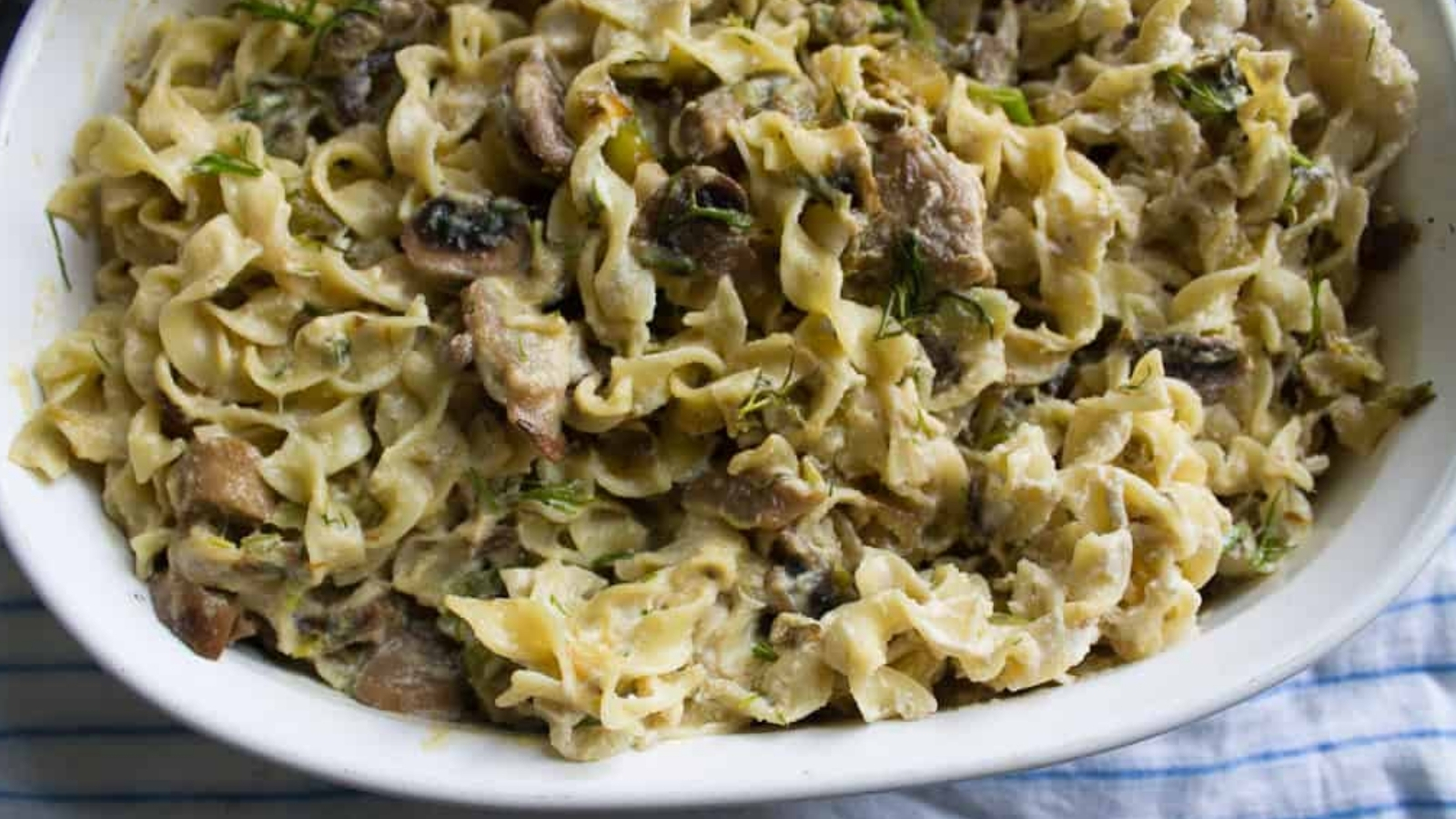 A white dish filled with creamy pasta topped with mushrooms and herbs is placed on a blue and white striped cloth.