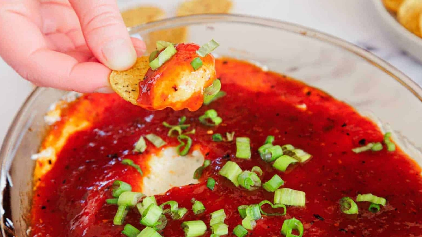 A hand dips a chip into a cheesy dip topped with red sauce and chopped green onions.