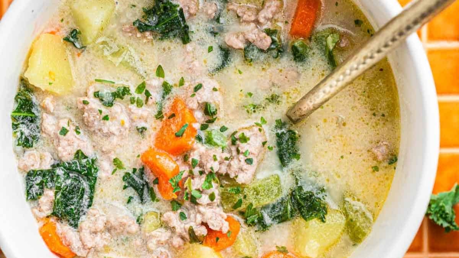 A bowl of creamy soup with sausage, kale, carrots, and potatoes, with a spoon inside, on an orange tiled surface.