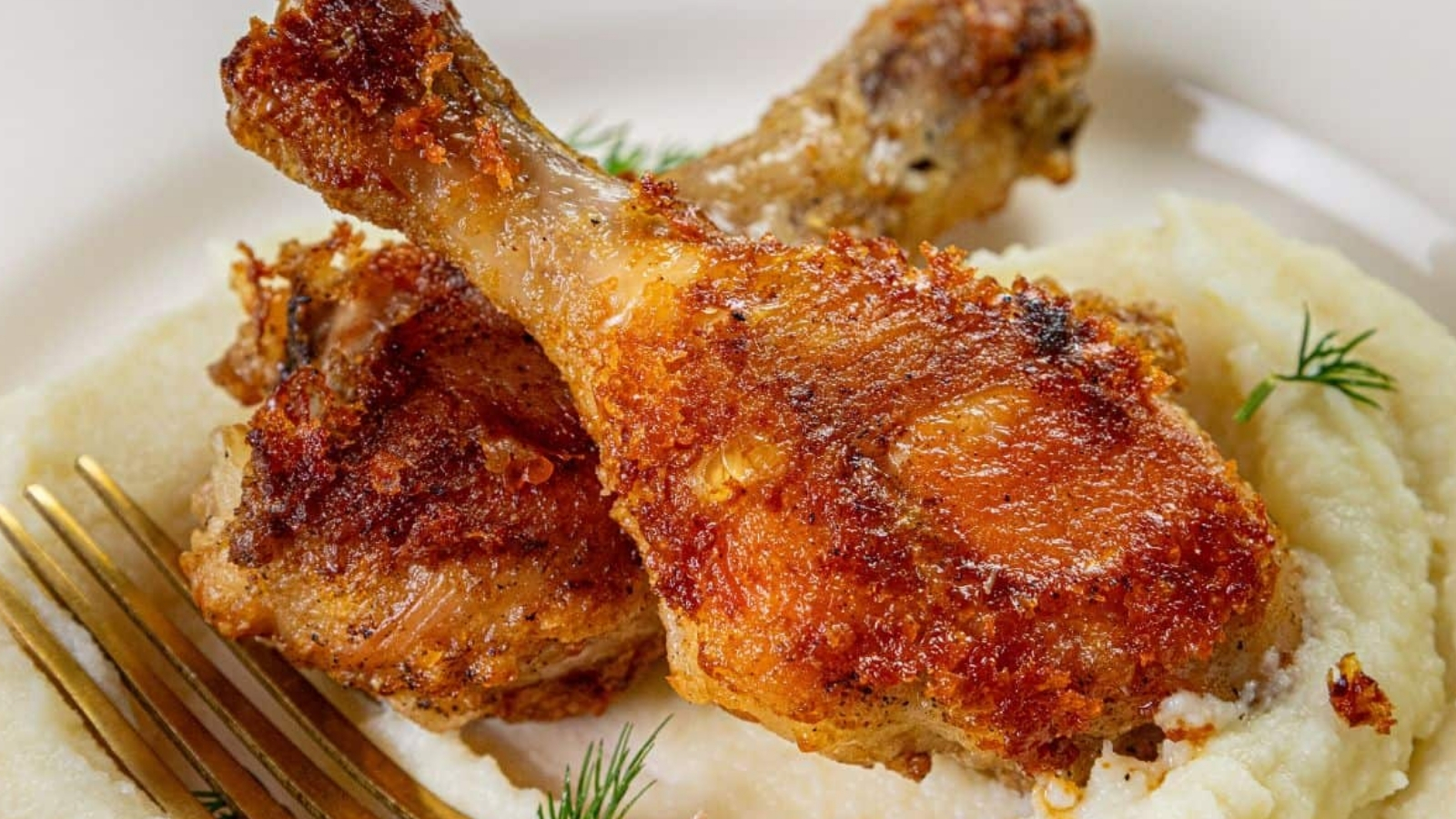Two crispy fried chicken drumsticks on a bed of creamy mashed potatoes, garnished with dill. A fork is placed beside the dish. The chicken is golden brown and the potatoes have a smooth texture.