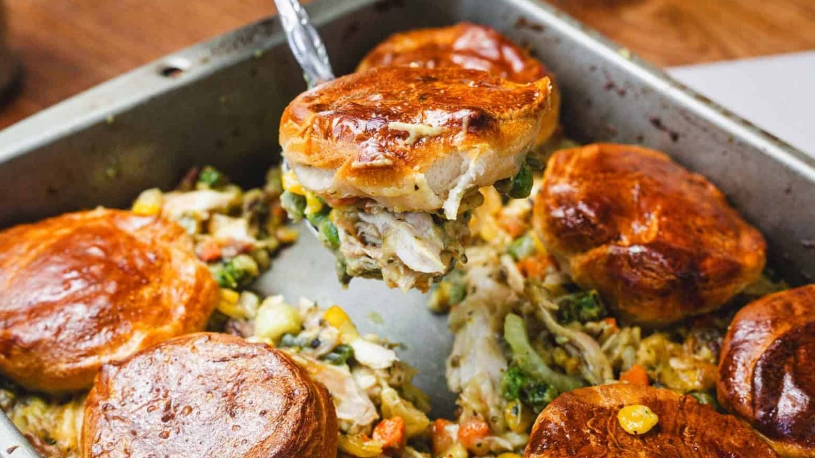 Golden biscuits on creamy chicken pot pie filling with vegetables in a baking pan, one biscuit being served.