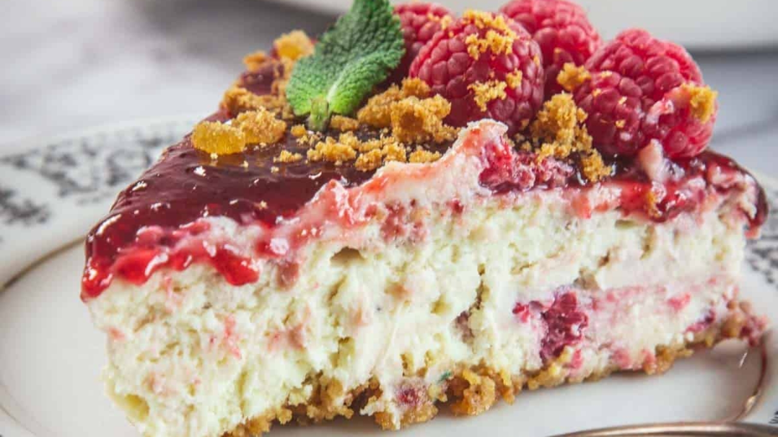 A slice of raspberry cheesecake topped with raspberries, mint leaves, and crumbled topping is placed on a decorative plate with a fork beside it.
