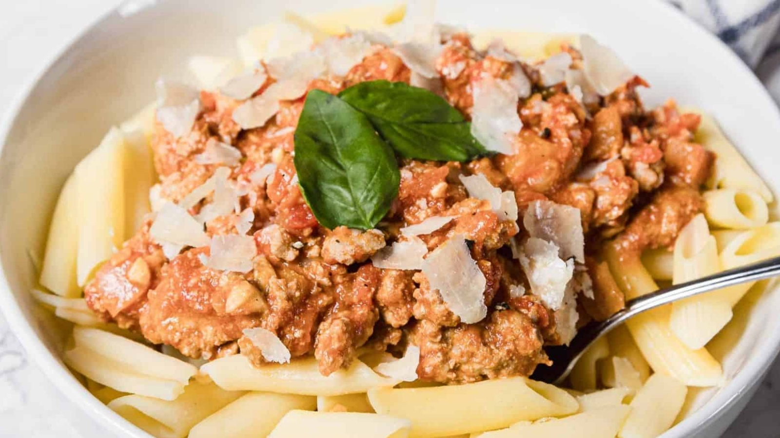 A white bowl filled with penne pasta topped with meat sauce, grated parmesan cheese, and fresh basil leaves, with a metal spoon resting on the side of the bowl.