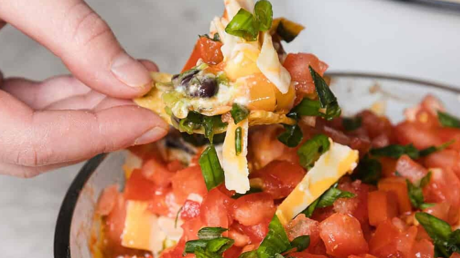 A hand dips a tortilla chip into a layered dip in a glass bowl. The dip consists of chopped tomatoes, green onions, cheese, beans, guacamole, and sour cream, arranged in visible layers. The bowl is on a marble surface, with more chips in the background.