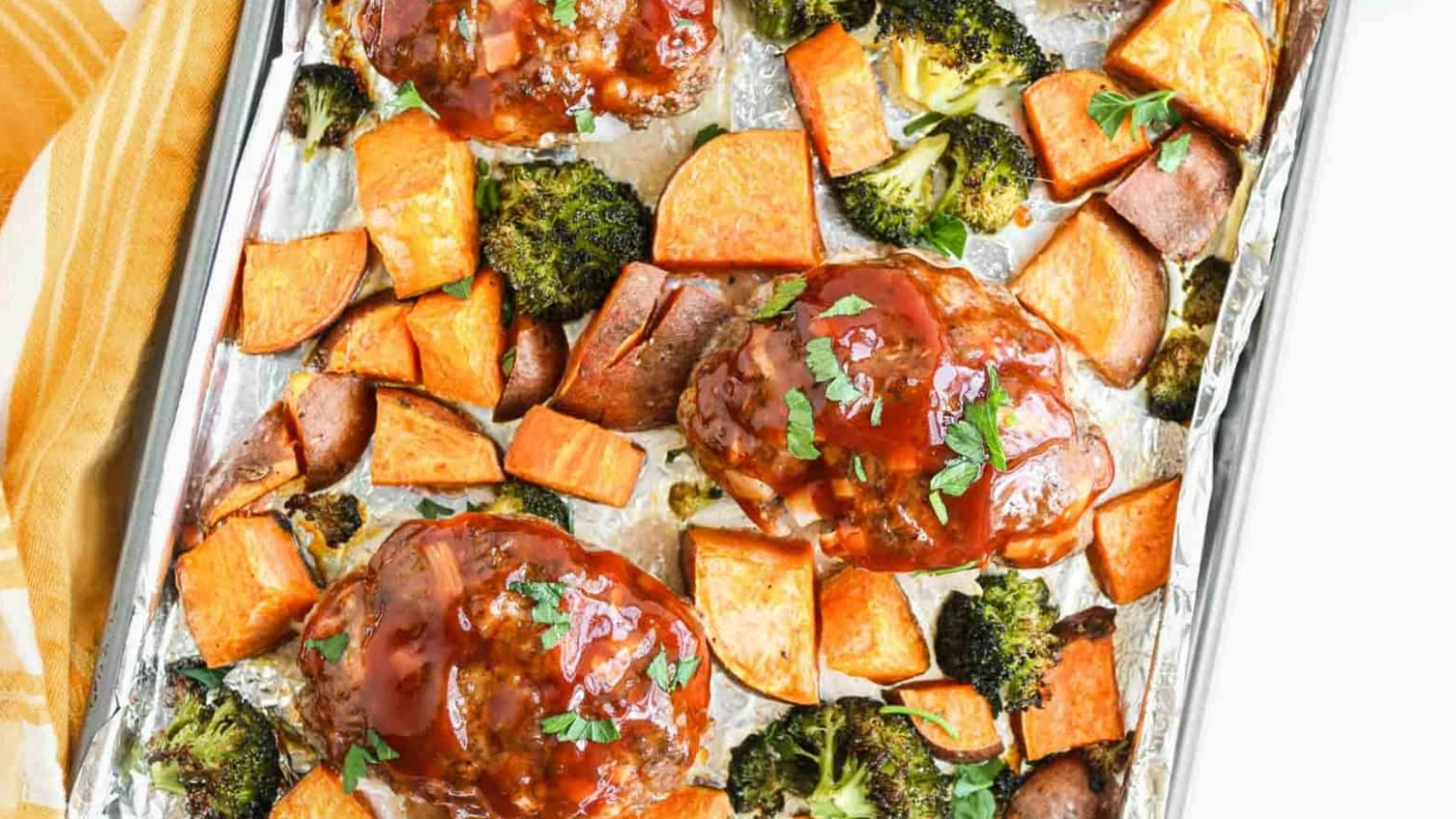 A baking sheet lined with foil holds three glazed meatloaf portions, surrounded by roasted sweet potato cubes and broccoli florets. A yellow and white towel is partially visible to the left.