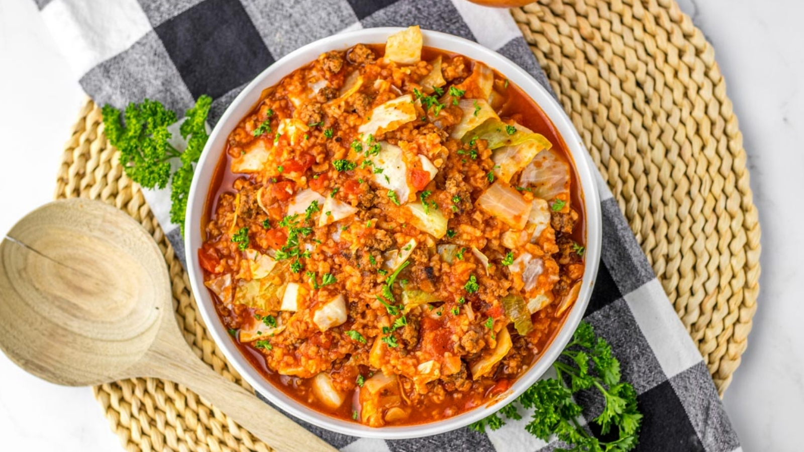 A bowl of stuffed cabbage roll soup with ground meat, cabbage, and tomato sauce, garnished with chopped parsley, sits on a woven placemat next to a wooden spoon.