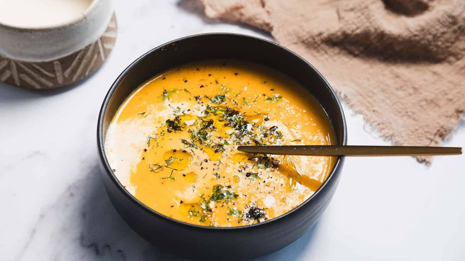 A black bowl filled with creamy orange soup, garnished with herbs, pepper, and a drizzle of oil. A gold spoon rests inside the bowl. A textured brown cloth and a cup are in the background.