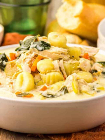 A bowl of creamy orange soup is placed on a black plate, garnished with a swirl of cream and a sprig of herb. A spoon rests inside the bowl. Small sprigs of thyme are scattered on the marble surface nearby.