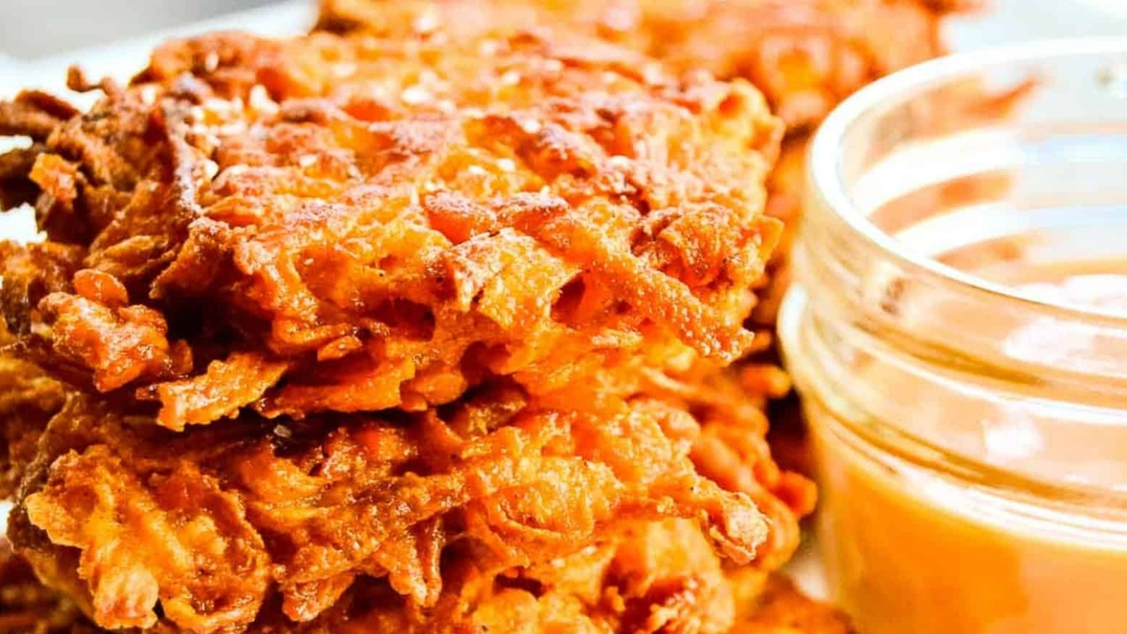A serving basket of sweet potato fritters next to a bowl of dipping sauce and a single serving on a plate.